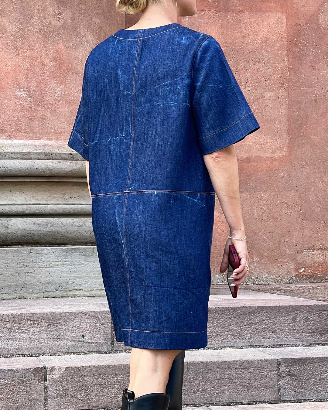 Person wearing a blue denim dress standing on stone steps with a red-brown wall in the background.