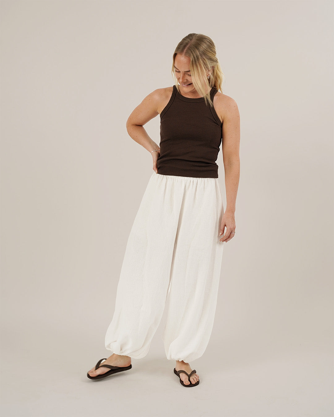 A woman wearing a brown muscle tank top and white bubble pants on a plain background