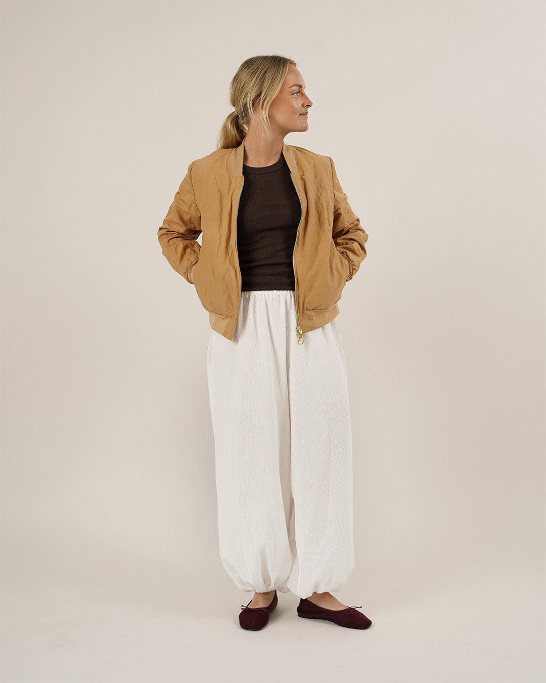 Woman wearing a tan jacket, brown top, white pants, and maroon shoes on a plain background