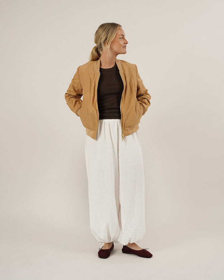 Woman wearing a tan jacket, brown top, white pants, and maroon shoes on a plain background