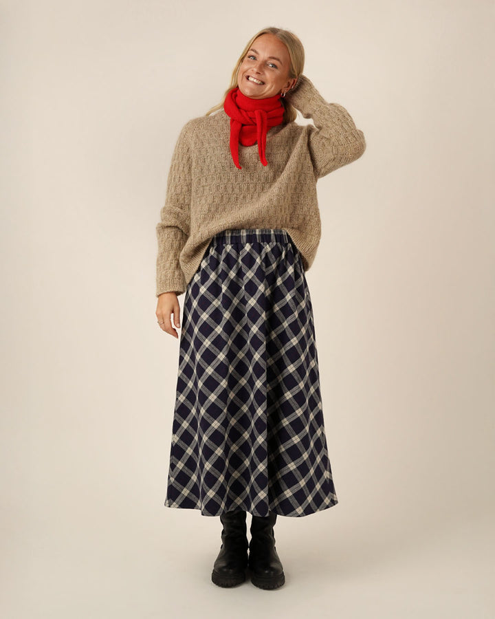 A woman wearing a Claude skirt sewing pattern with a beige sweater, red scarf,  on a plain background
