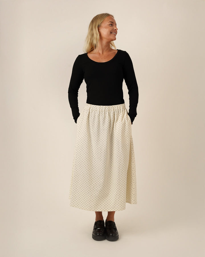 A woman wearing a black wardrobe Builder T-shirt sewing pattern, and a white polka dot Claude skirt sewing patternon a beige background