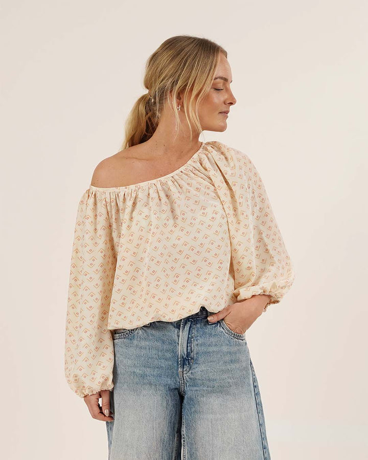 A woman wearing a floral moira blouse off-shoulder top and jeans on a plain background
