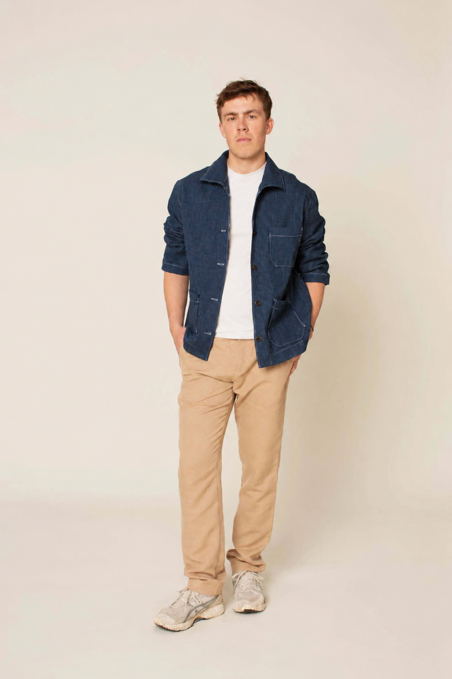 A man wearing a dark blue textured jacket over a white t-shirt, paired with beige trousers and light gray sneakers, standing against a plain light beige background.