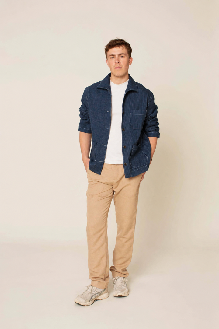 A man wearing a dark blue textured jacket over a white t-shirt, paired with beige trousers and light gray sneakers, standing against a plain light beige background.