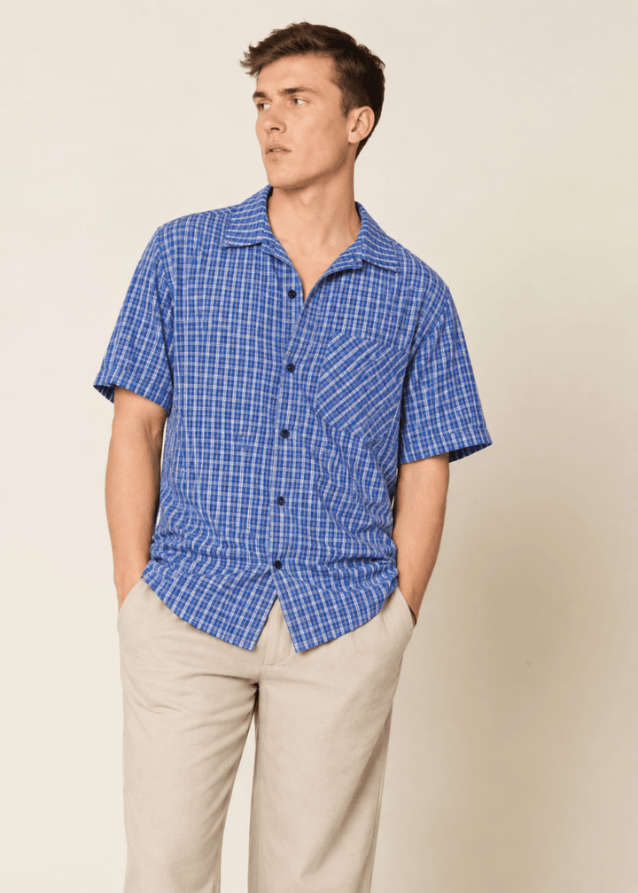 A man wearing a blue and white checkered Tropical Shirt Sewing Pattern and beige trousers, standing against a plain beige background.