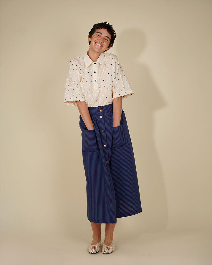 A woman modeling a cream short-sleeve button-up shirt with small black polka dots, paired with a navy blue wrap skirt featuring gold buttons, against a solid beige studio background.