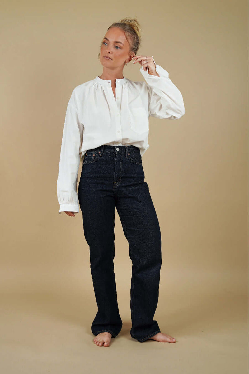 Woman wearing a white blouse and dark jeans against a beige background