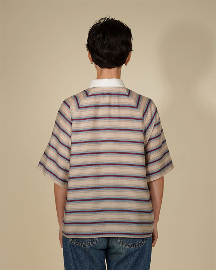 A person viewed from the back wearing a short-sleeve collared shirt with horizontal beige, white, and navy stripes, paired with blue jeans, against a plain beige studio background.