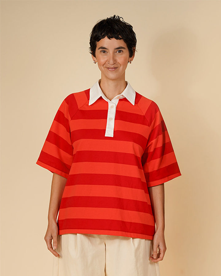 A model wearing a red and orange horizontally striped short-sleeve polo shirt with a white collar, paired with cream pants, against a solid beige background.