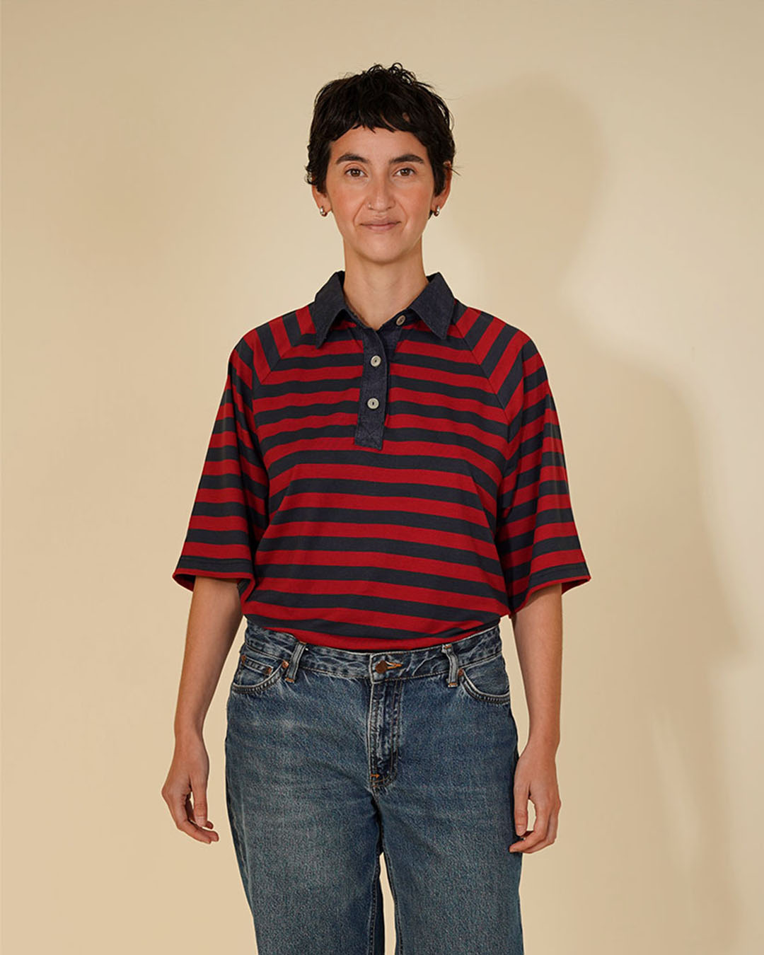 A person wearing a short-sleeve polo shirt with horizontal red and navy stripes, paired with blue jeans, standing against a plain beige studio background.