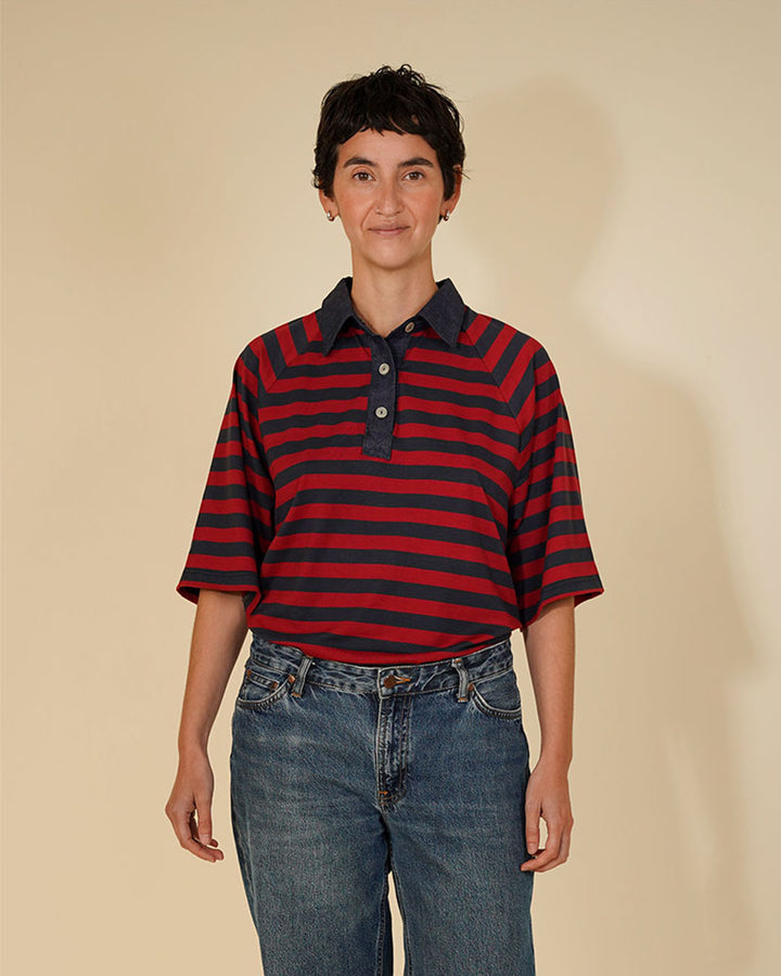 A person wearing a short-sleeve polo shirt with horizontal red and navy stripes, paired with blue jeans, standing against a plain beige studio background.
