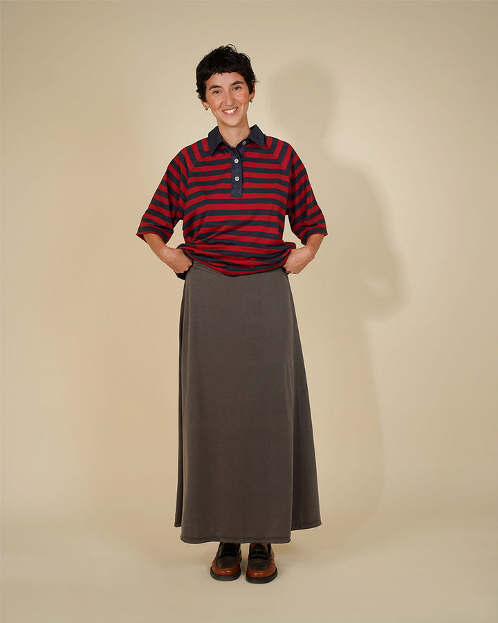 A person standing against a plain beige studio background, wearing a short-sleeve red and navy striped polo shirt with a dark collar and buttons, paired with a long dark gray skirt and dark brown shoes.