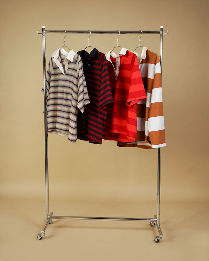 Five short-sleeve polo shirts in various stripe and solid patterns, hanging on a silver metal rolling clothing rack against a solid beige background.