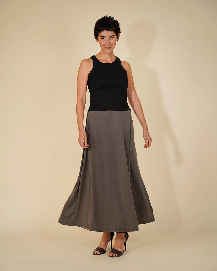 A woman modeling a sleeveless black ribbed tank top paired with a high-waisted, flowing taupe-gray maxi skirt, styled with black heeled sandals against a warm beige studio background.