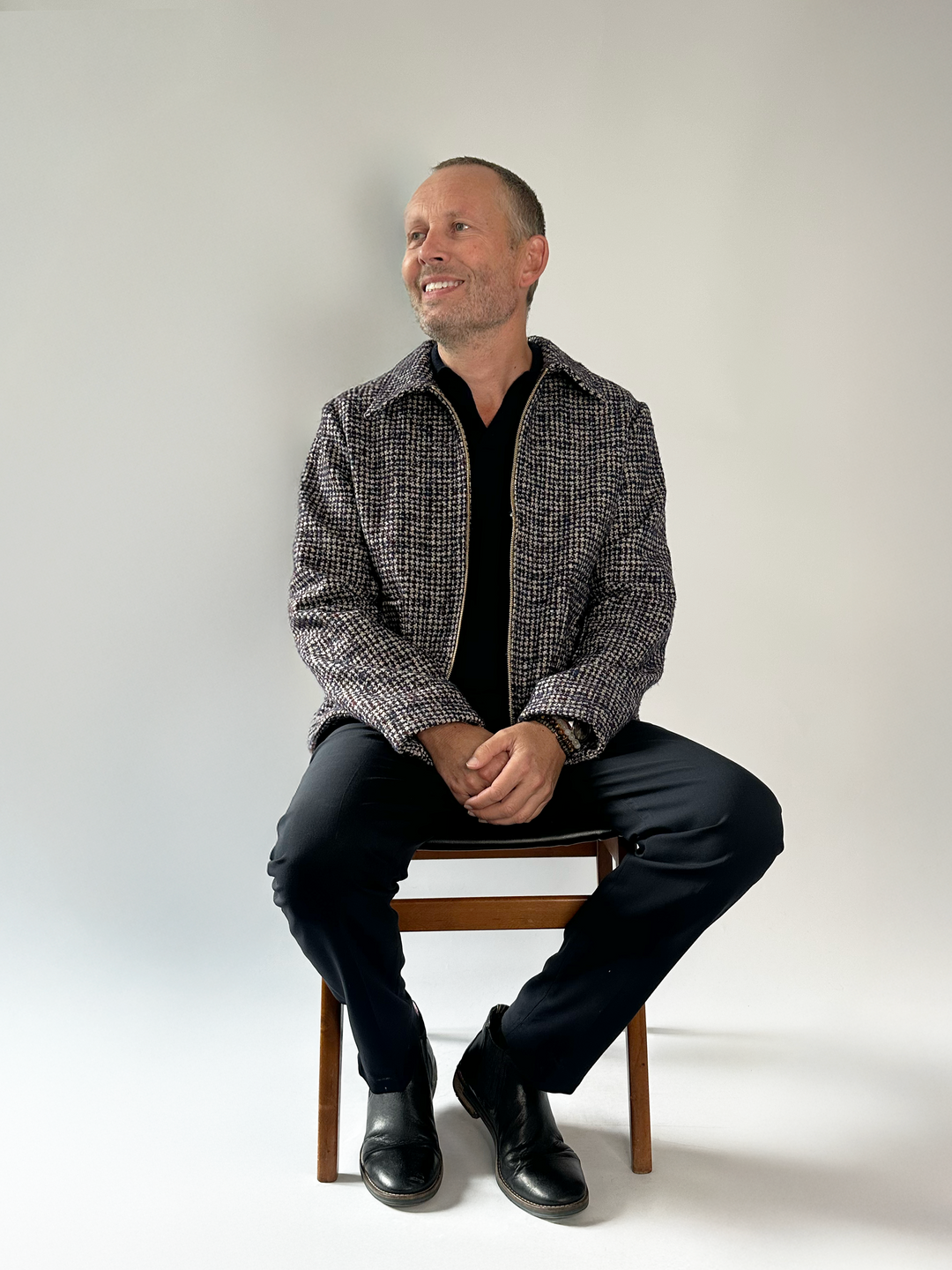 Man sitting on a chair wearing a checkered jacket and black pants against a white background