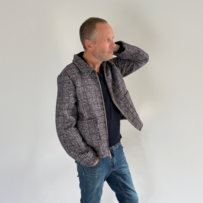 Man wearing a checkered jacket, dark shirt, blue jeans, and white sneakers on a plain background
