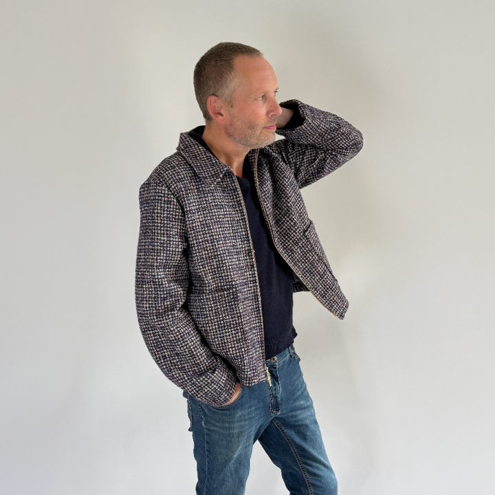 Man wearing a checkered jacket, dark shirt, blue jeans, and white sneakers on a plain background