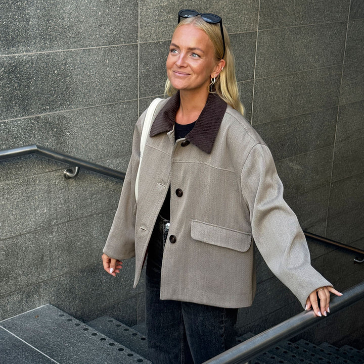 Woman in a beige jacket standing on a staircase against a gray wall.