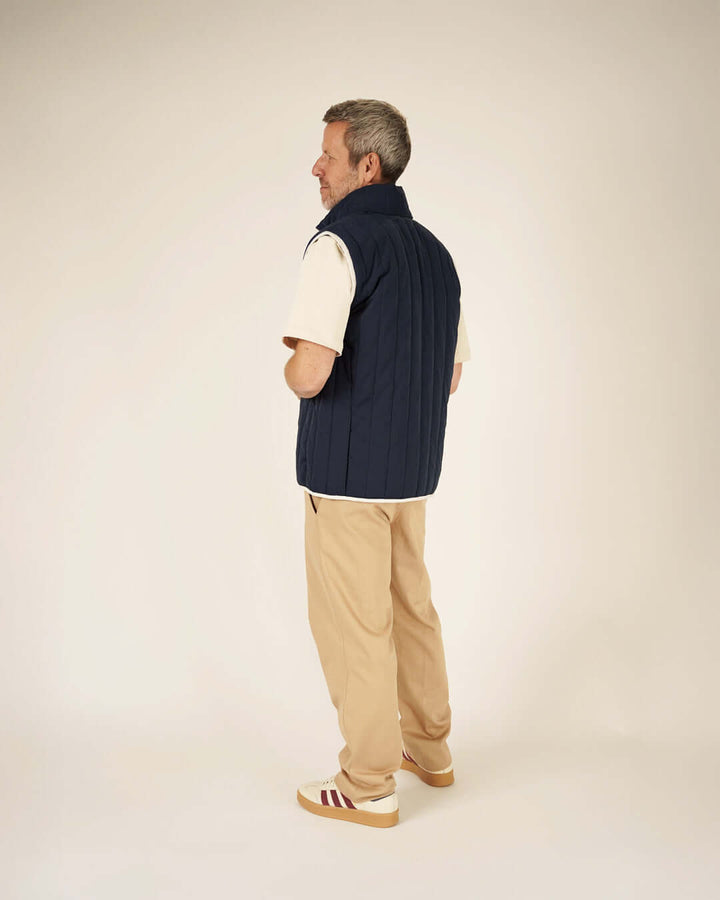 Man wearing a navy vest over a beige shirt and tan pants on a plain background