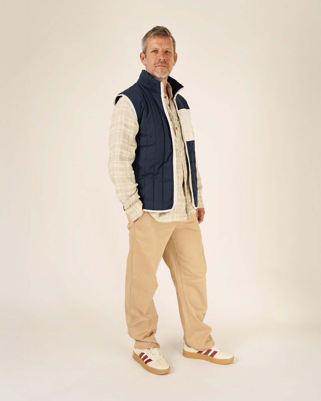 Man wearing a blue vest over a plaid shirt with beige pants and shoes on a white background