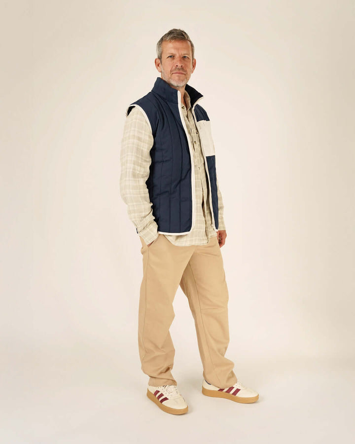 Man wearing a blue vest over a plaid shirt with beige pants and shoes on a white background