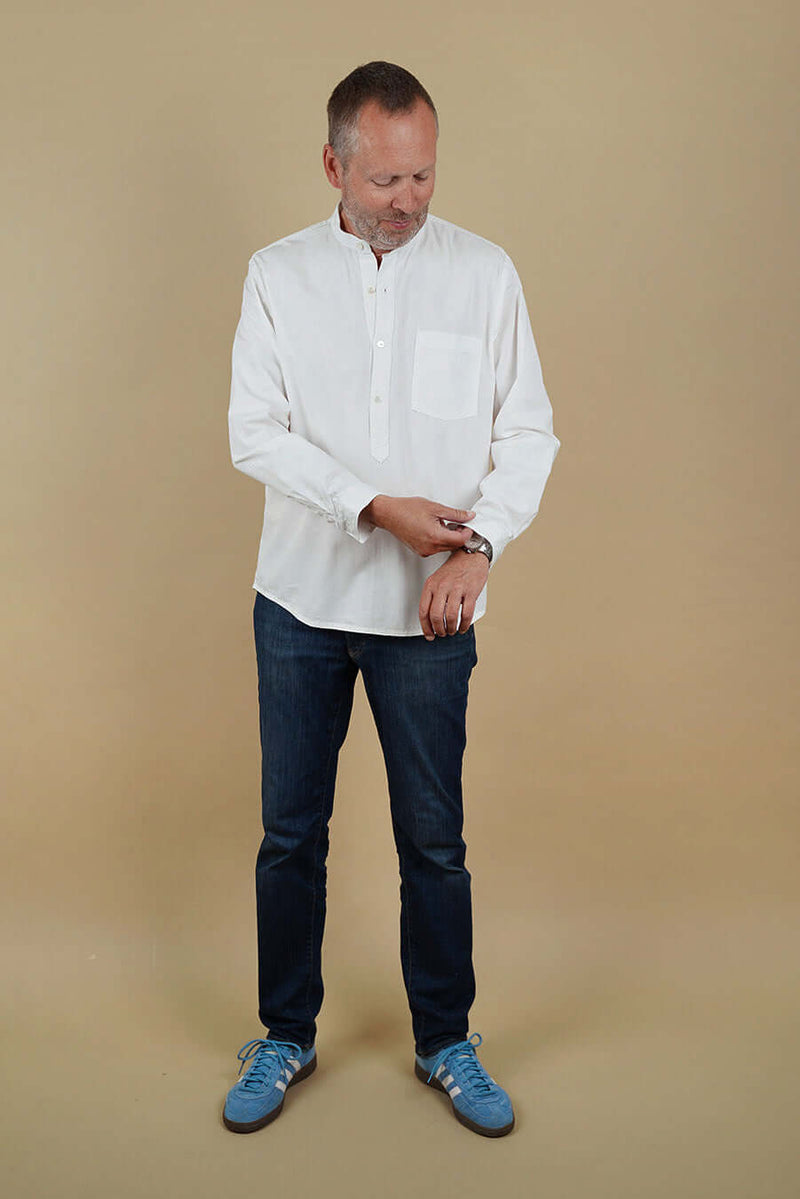 A man wearing a white tunic, dark jeans, and blue shoes on a beige background