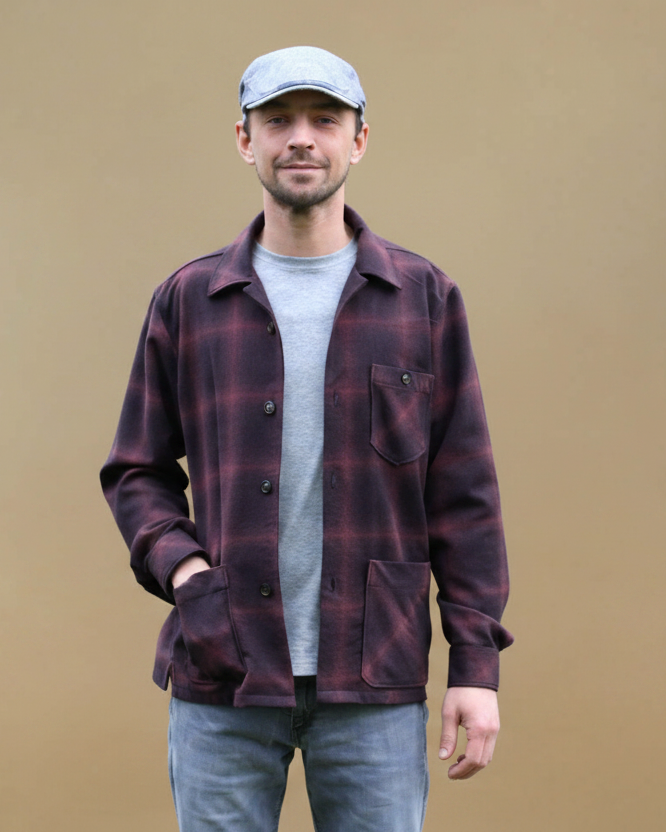 A man wearing a dark red and black plaid overshirt jacket over a light gray t-shirt, blue jeans, and a gray cap, standing against a solid tan background.