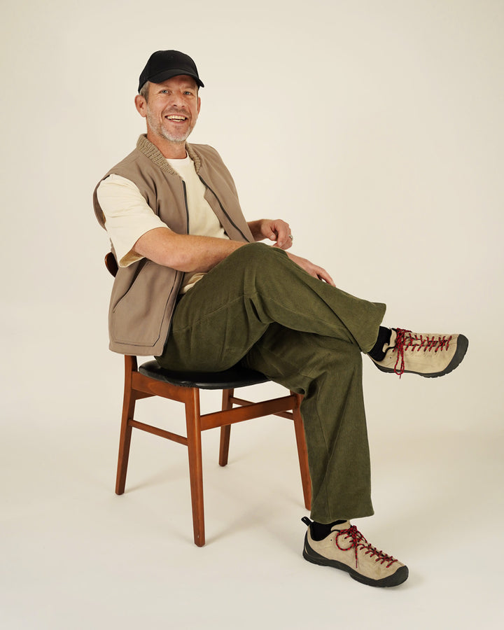 Man sitting on a chair wearing a vest, shirt, pants, and shoes against a plain background