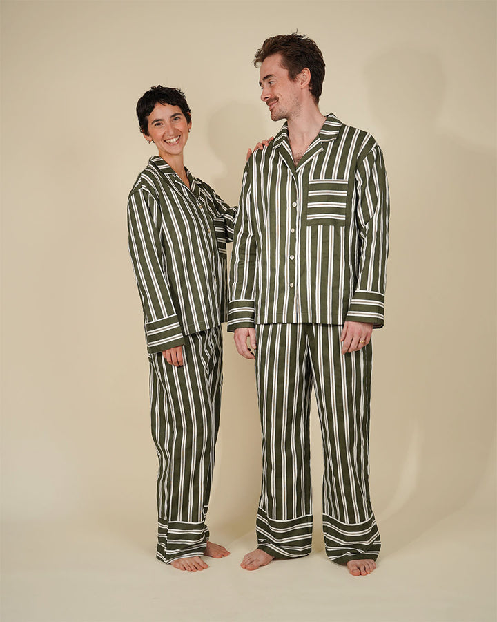 A man and a woman wearing matching striped pajama sets, standing barefoot against a plain beige studio background. The pajamas feature vertical stripes in dark green and off-white.