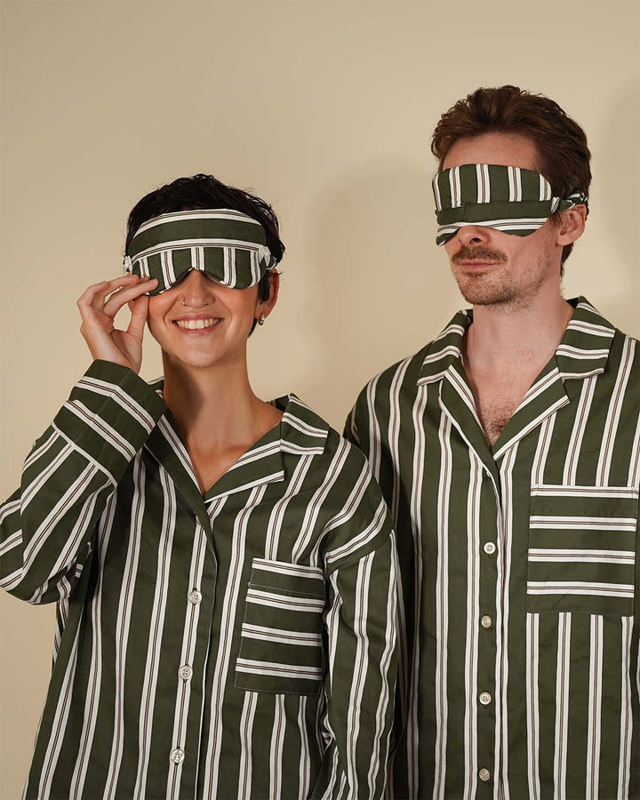 A man and a woman wearing matching green and white vertically striped pajamas and blindfold sleep masks, posing against a solid beige background.