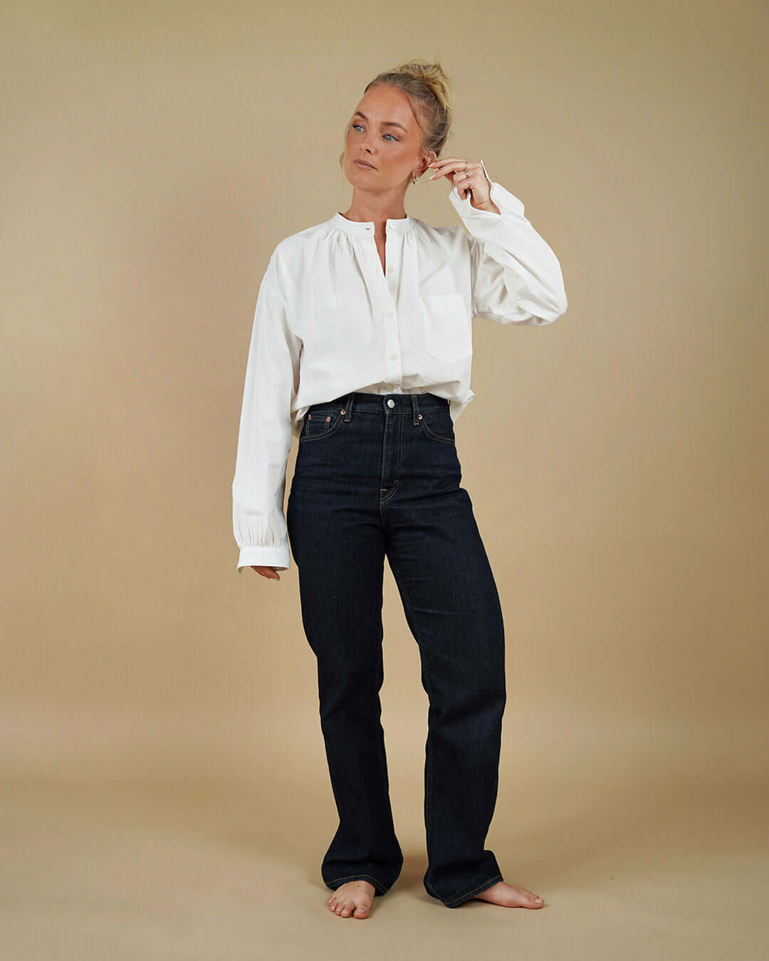 A woman stands barefoot against a plain beige studio background, wearing a loose white long-sleeve blouse and dark navy straight-leg jeans.