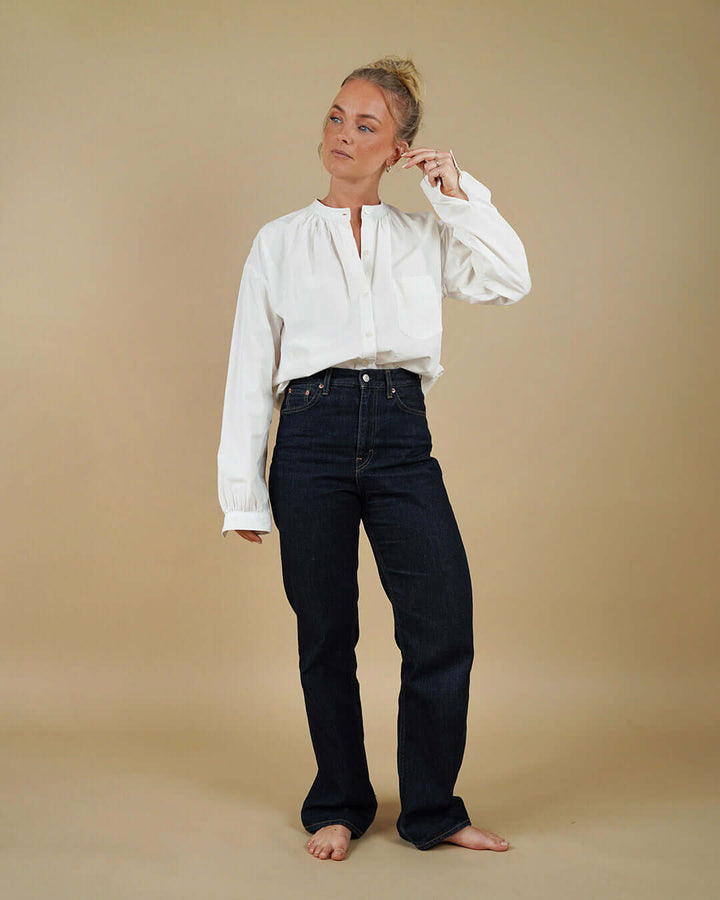 A woman stands barefoot against a plain beige studio background, wearing a loose white long-sleeve blouse and dark navy straight-leg jeans.