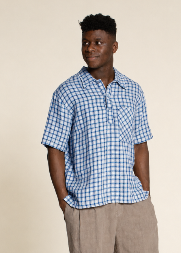 A man wearing a blue and white checkered short-sleeve button-up shirt with beige trousers, standing against a solid beige background.