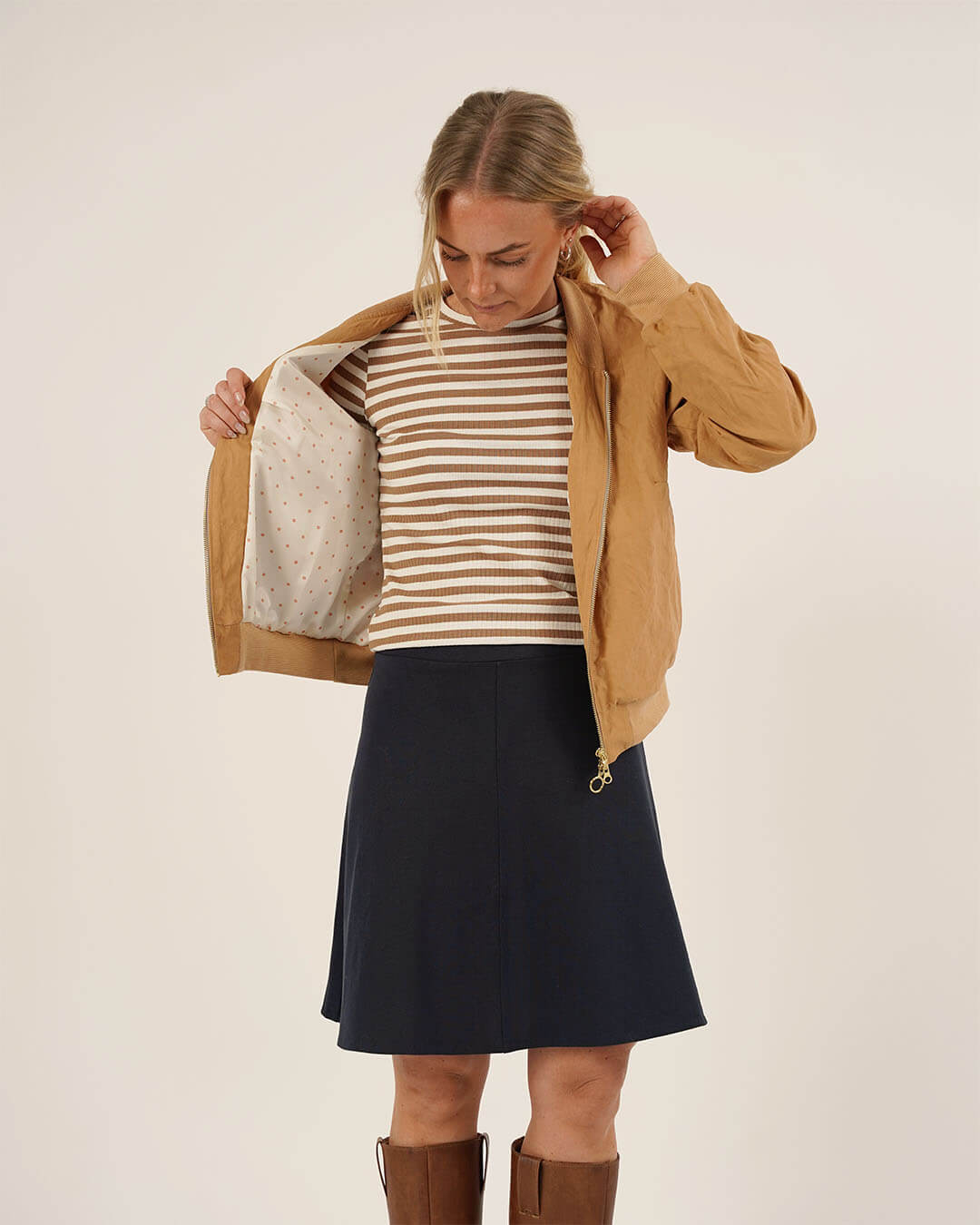 Woman wearing the Amelia Bomber Jacket sewing pattern in camel fabric over a striped knit top and paired with the Linea Skirt sewing pattern in navy fabric, showing the jacket lining