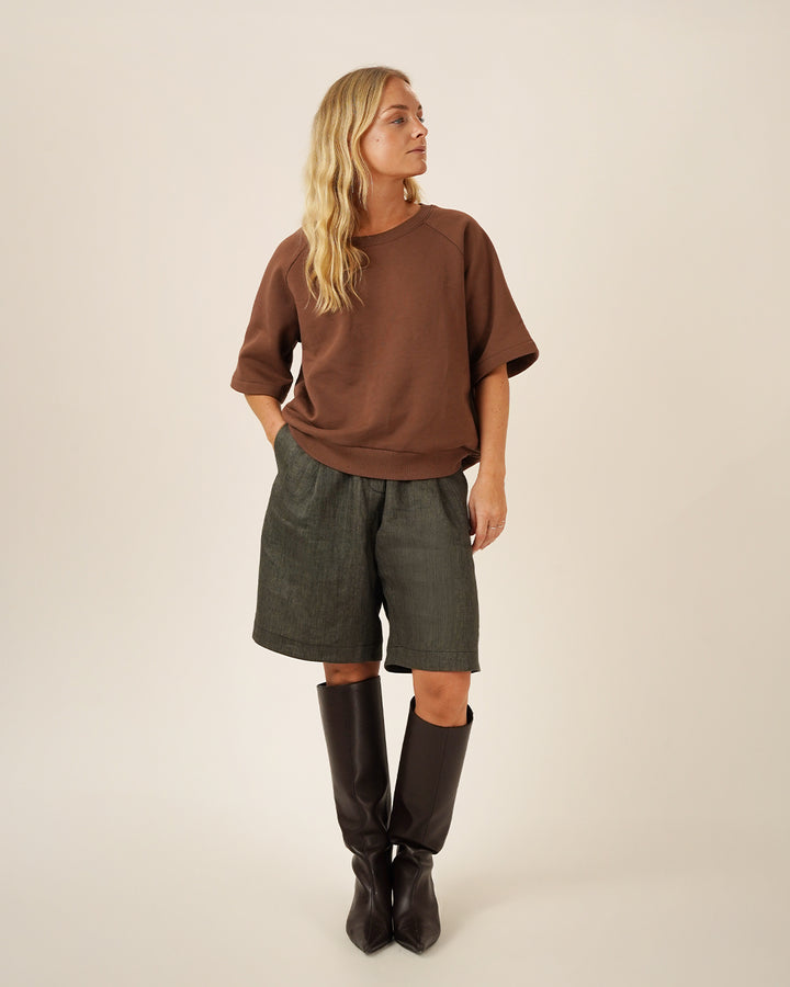 Woman wearing the Boxy Raglan sewing pattern in brown sweatshirt fabric with elbow-length sleeves, styled with dark Bermuda shorts and knee-high boots