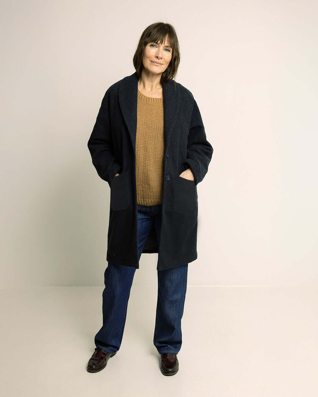Woman wearing the Cocoon Coat sewing pattern in dark wool fabric over a camel knit sweater and jeans, front view with hands in patch pockets