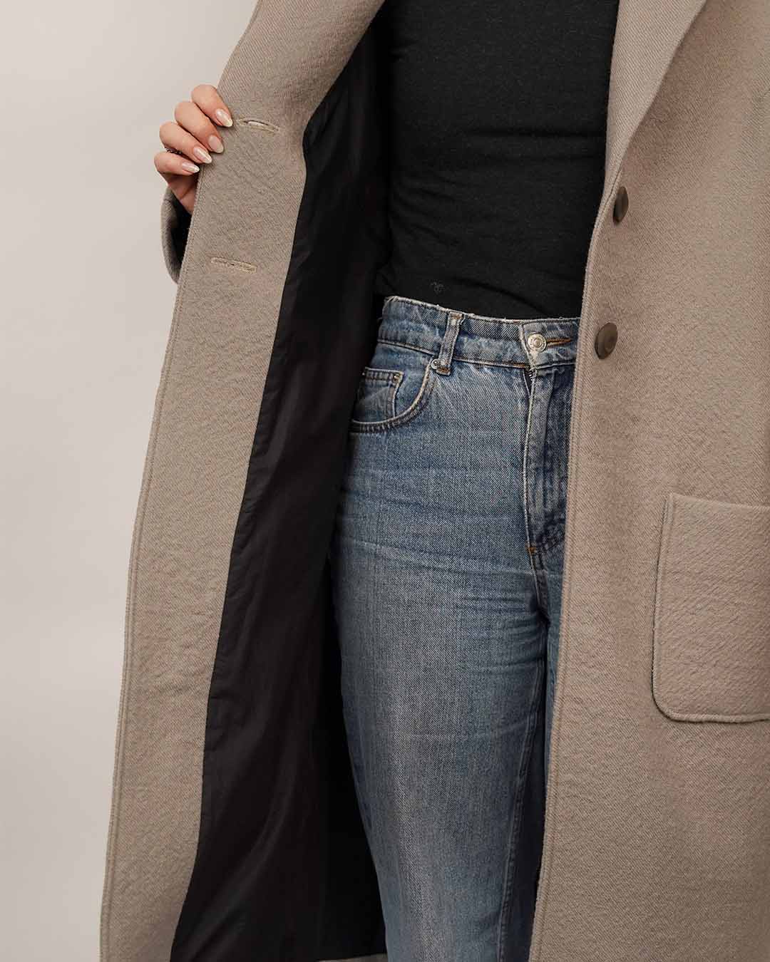 Close-up of the Cocoon Coat sewing pattern showing the coat lining, button closure and patch pocket details, layered over a black Trudy Turtleneck and blue jeans