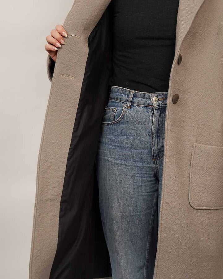 Close-up of the Cocoon Coat sewing pattern showing the coat lining, button closure and patch pocket details, layered over a black Trudy Turtleneck and blue jeans