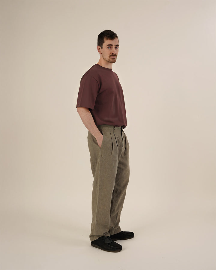 Man wearing the Men's Pleated Pants and Big T-shirt sewing patterns, shown from a three-quarter side view in taupe pleated trousers and a dark mauve oversized T-shirt