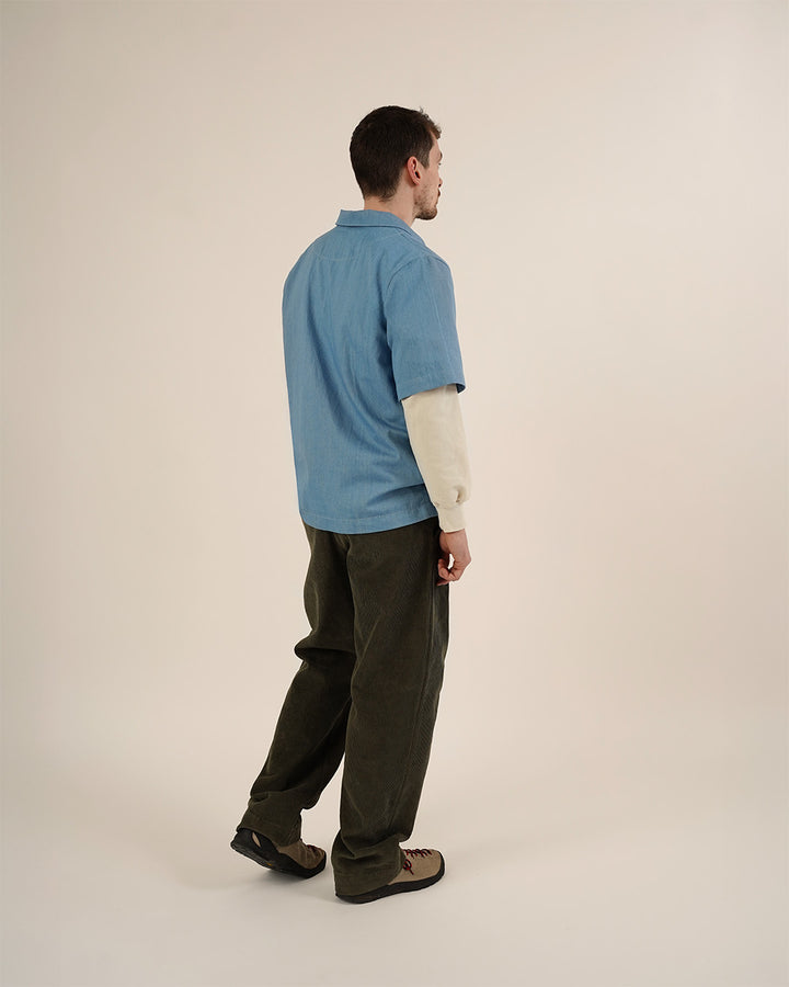 Man wearing the Men's Zip Shirt and Barrel Pants sewing patterns, shown from a three-quarter back view in a light blue short-sleeve shirt layered over a cream long-sleeve top with dark barrel-leg trousers