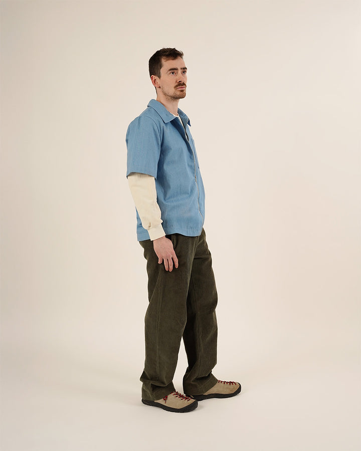 Man wearing the Men's Zip Shirt and Barrel Pants sewing patterns, shown from a three-quarter front view in a light blue short-sleeve shirt layered over a cream long-sleeve top with dark barrel-leg trousers